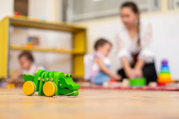Drewniany krokodyl na podłodze żłobka. W tle opiekunka z dziećmi. Ciekawe, ile zarabia opiekun w żłobku państwowym za taką pracę.