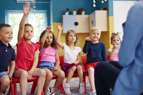 Dzieci na czerwonych krzesłach podnoszą ręce, odpowiadając na pytanie o to, ile lat się chodzi do przedszkola.