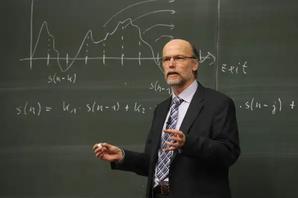 Profesor w garniturze tłumaczy równania na tablicy. Zastanawiamy się, ile godzin ma nauczyciel na etacie, gdy widzi się jego zaangażowanie.